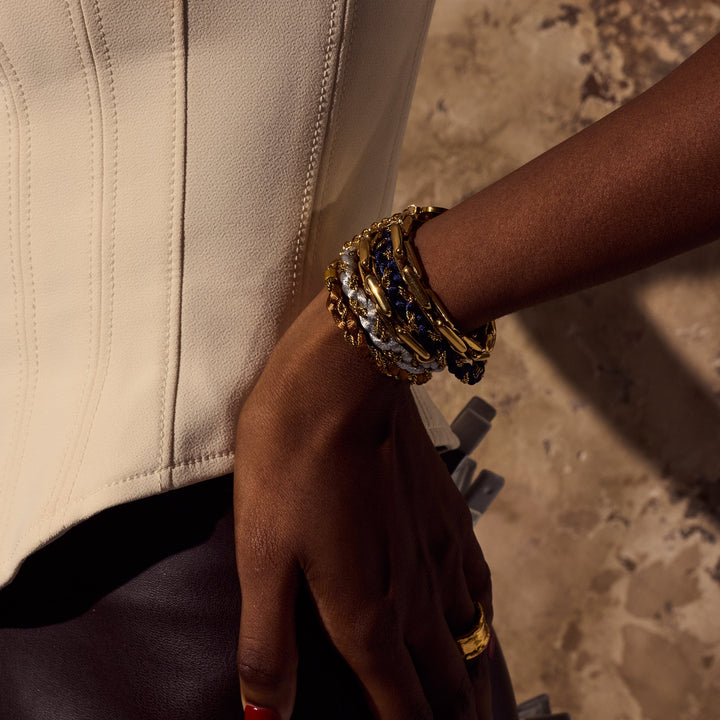 A close-up of a person's hand and wrist wearing a stack of chunky gold bracelets, some with blue inlay details, paired with a cream-colored leather jacket.