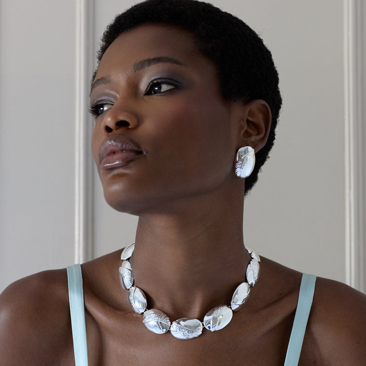 A woman with short dark hair is shown from the chest up against a light paneled wall, wearing a chunky white shell necklace and matching white shell earrings.
